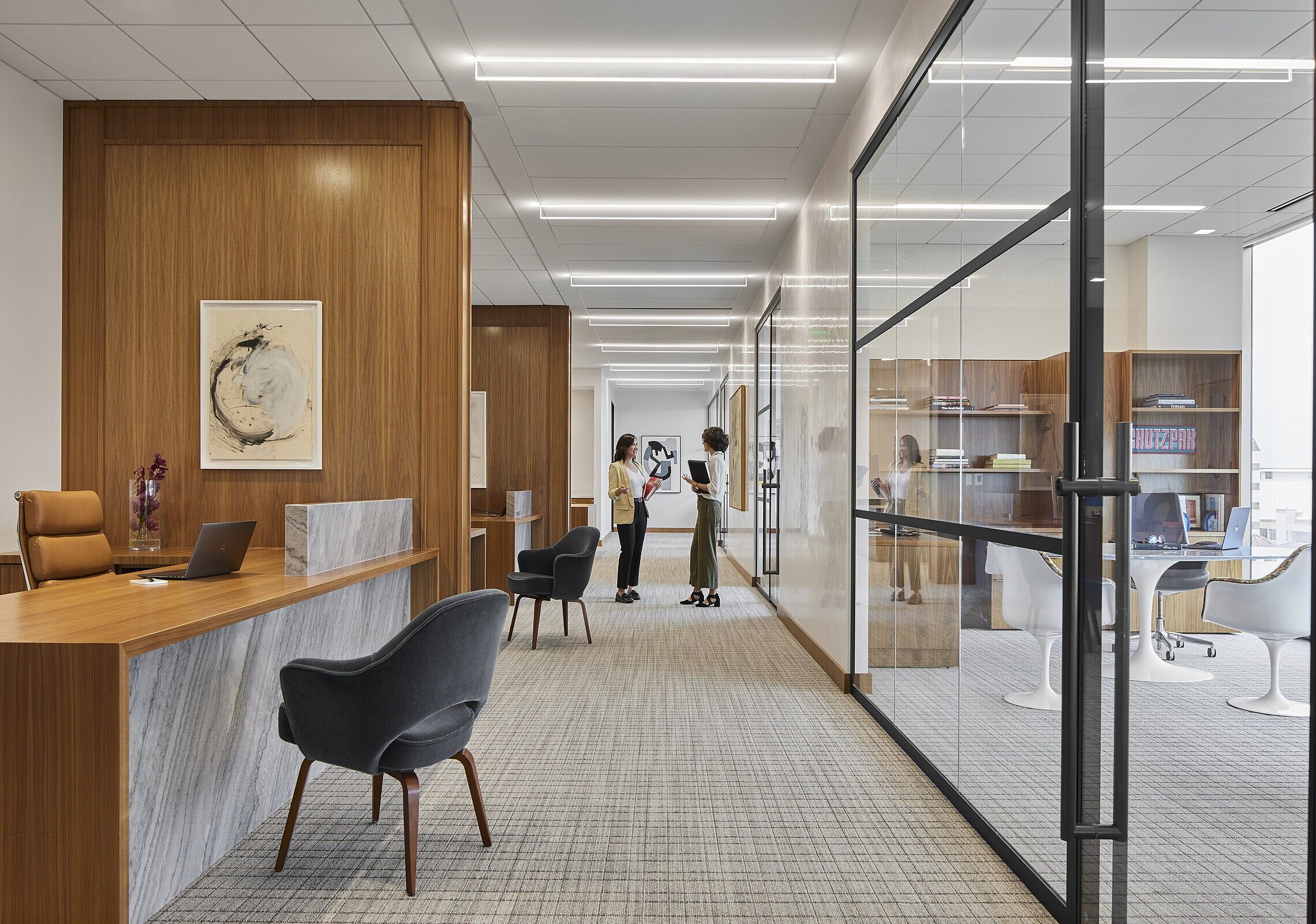 Desks with a guest chairs on the left down a hallway with private offices behind glass doors to the right
